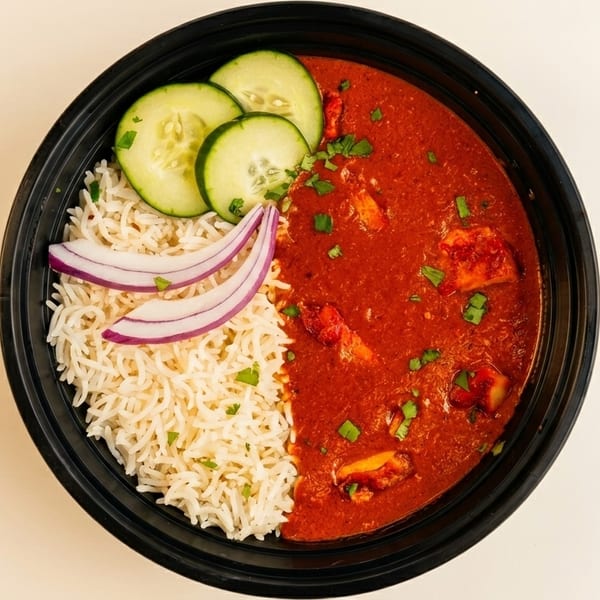 Overhead view of a black bowl with chicken tikka masala, rice, cucumber slices, and red onion.